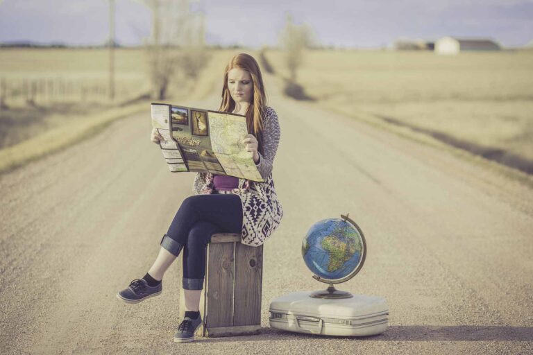 The photo shows a girl sitting in the middle of a deserted street looking at a map she holds in her hands. Next to her is a suitcase and a globe.