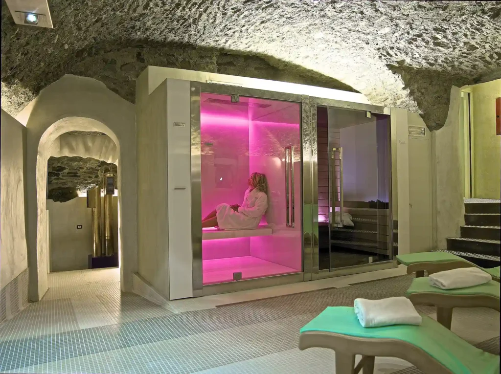 L'immagine mostra l'area SPA dell' hotel caratterizzata a un soffitto in pietra. Si intravedono tre lettini. DI fronte un bagno turco, Dentro la sauna c'è una ragazza che si rilassa.
