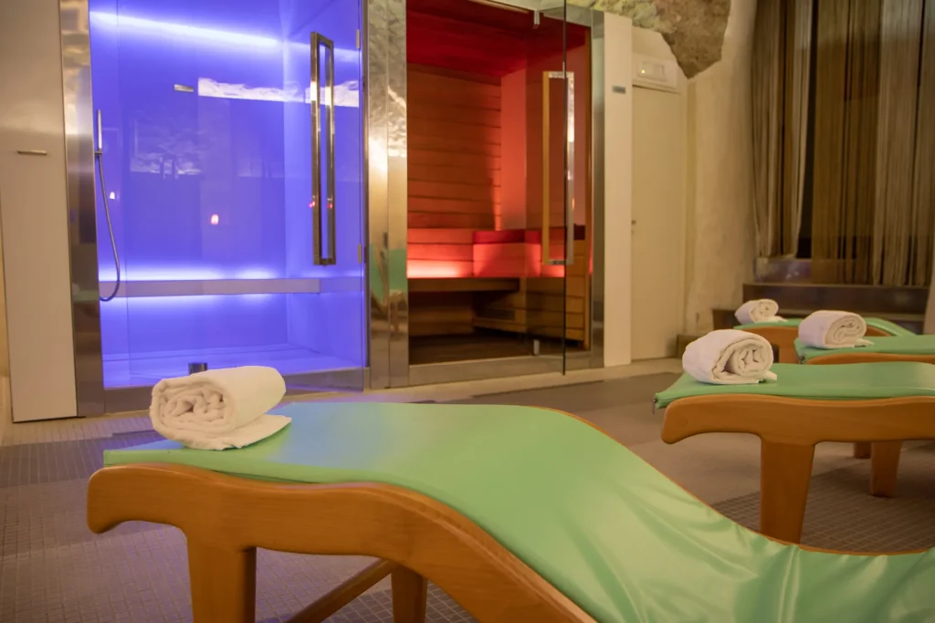Nell' immagine in primo piano quattro lettini dove potersi rilassare. Sullo sfondo il bagno turco e la sauna illuminati con luci soffuse.